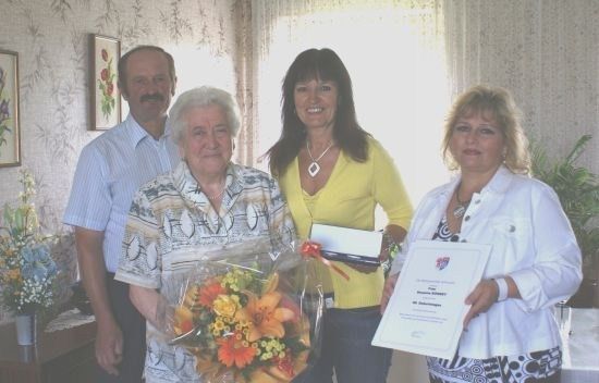 Eine ältere Frau mit einem Blumenstrauß steht zwischen zwei Frauen, eine hält ein Zertifikat und die andere ein gerahmtes Dokument. Hinter ihnen steht ein Mann neben einer Vase mit Blumen.