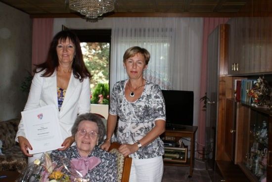 Drei Frauen in einem Wohnzimmer, eine hält ein Zertifikat und Blumen, eine andere steht neben einer älteren Frau im Stuhl, und eine dritte Frau steht hinter ihnen.