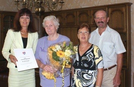 Vier Menschen stehen in einem Raum, einer hält ein Zertifikat und Blumen, ein anderer einen Blumenstrauß und zwei andere stehen neben ihnen.
