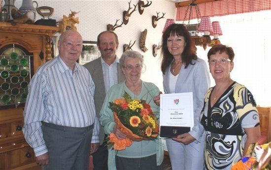 Fünf Menschen stehen in einem Raum zusammen, eine Frau hält einen Blumenstrauß und ein Zertifikat in der Hand. Die Frau trägt eine Brille und eine Halskette. Hinter ihr stehen zwei Männer und zwei Frauen. Der Raum hat einen Holzschrank mit einer Glastür und eine Lampe, die von der Decke hängt.