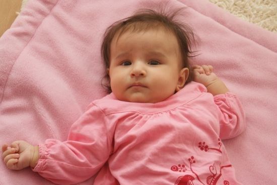 Ein Babymädchen mit kurzen Haaren liegt auf einer rosa Decke und trägt ein rosa Langarmshirt mit Blumenmuster. Sie schaut nach links.