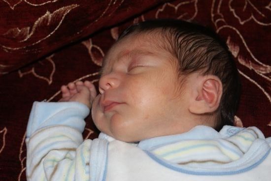 Ein schlafendes Baby liegt auf einem gemusterten roten Stoff und trägt ein weiß-blau gestreiftes Overall. Die Augen des Babys sind geschlossen und die Hände leicht erhoben.