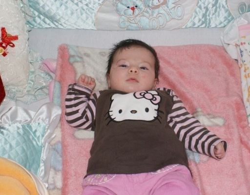 Ein Babymädchen liegt auf einer rosa Decke mit einem Hello Kitty Shirt und rosa Hosen. Sie ist von bunten Kuscheltieren und Decken umgeben.
