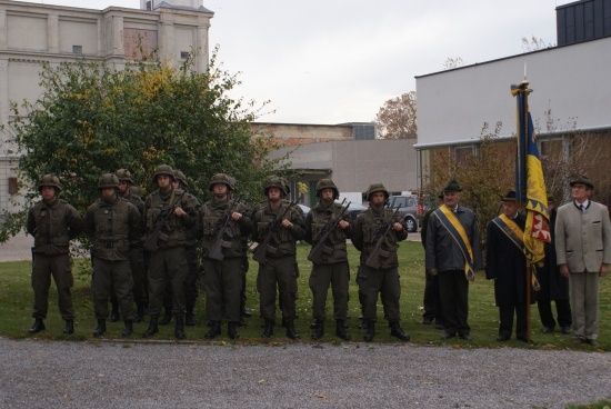 Eine Gruppe von Soldaten in Militäruniformen steht in Formation auf einem Rasen mit Gebäuden im Hintergrund.