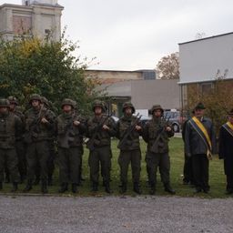 Eine Gruppe von Soldaten in Militäruniformen steht in Formation auf einem Rasen mit Gebäuden im Hintergrund.