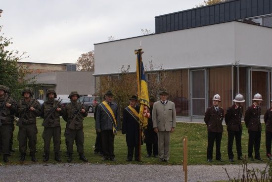 Eine Gruppe von Menschen in Militäruniformen steht vor einem Gebäude mit einem Fahnenmast. Einige halten Gewehre, andere tragen Mützen. Das Gebäude hat Glasfenster und ein graues Dach.
