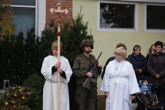 Zwei Personen in weißen Gewändern, eine mit einem Kreuz, stehen neben einer Person in Militäruniform mit einer Waffe, mit mehreren Leuten dahinter vor einem Gebäude.