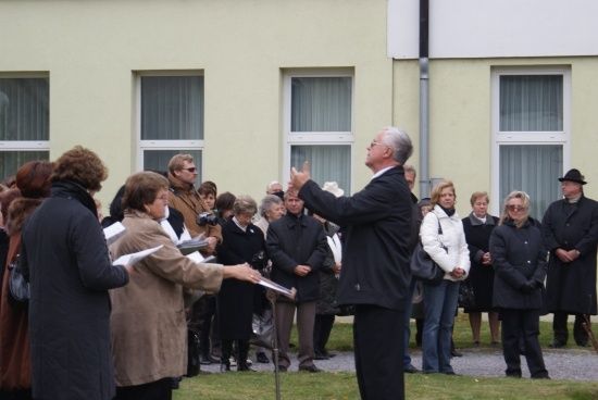 Eine Gruppe von Menschen steht vor einem Gebäude, wobei ein Mann im Anzug gestikuliert, während er spricht. Einige Leute halten Papiere in der Hand, während andere aufmerksam zuhören.