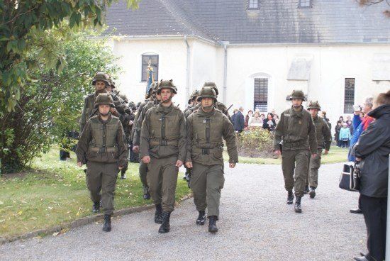 Eine Gruppe von Soldaten in Militäruniformen marschiert in einer Reihe auf einem Kiesweg vor einem Gebäude, wobei Zuschauer an der Seite stehen.