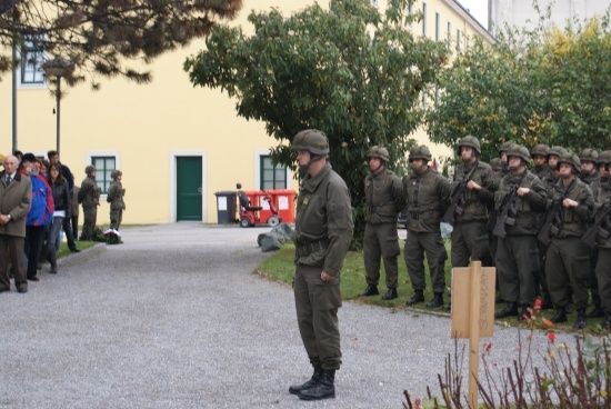 Eine Gruppe von Soldaten in Uniform steht in Formation vor einem Gebäude, wobei ein Mann im Vordergrund sie ansieht.