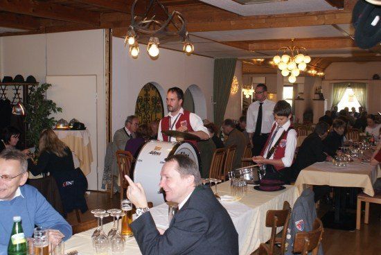 Eine Gruppe von Menschen versammelt sich in einem Restaurant, einige spielen Instrumente. Ein Mann zeigt auf einen anderen Mann, der an einem Tisch sitzt.