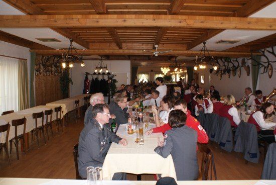 Eine Gruppe von Menschen in formeller Kleidung sitzt um einen langen Tisch in einem Bankettsaal, einige essen, andere unterhalten sich. Der Saal hat Holzbalken und Kronleuchter.