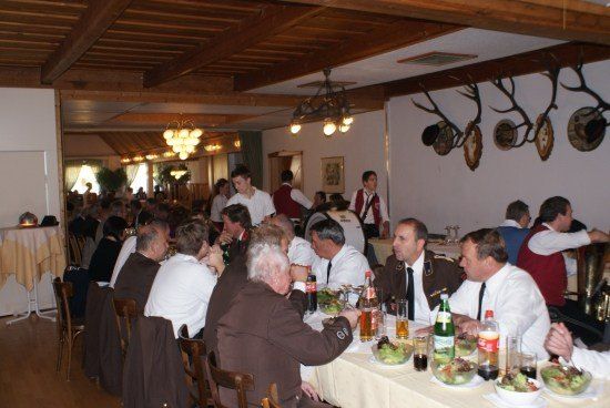 Eine Gruppe von Männern in Uniformen und Krawatten sitzt um einen Esstisch mit Essen und Getränken. Sie befinden sich in einem Raum mit Holzbalken und Geweihen an der Wand.