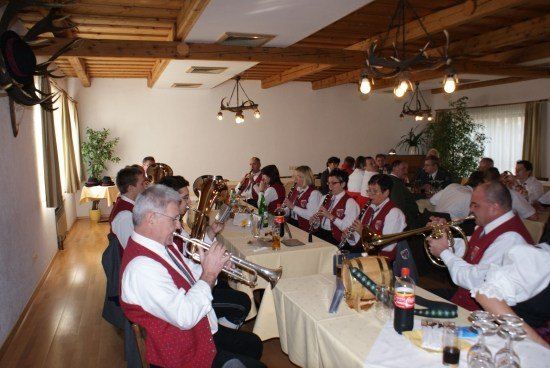 Eine Gruppe von Musikern in traditioneller Kleidung spielt verschiedene Instrumente in einem Raum mit Holzbalken und Kronleuchtern. Flaschen und Gläser stehen auf den Tischen.
