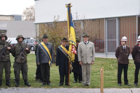 Eine Gruppe von Menschen in Militäruniformen und Mützen steht auf einem Rasen, möglicherweise für eine Zeremonie, mit einer Flagge in der Mitte. Dahinter befindet sich ein Gebäude mit Glasfenstern.