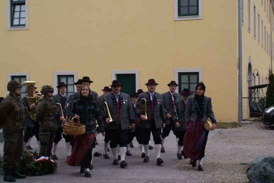 Eine Gruppe von Menschen in traditioneller Kleidung, darunter einige in Militäruniformen, marschiert vor einem gelben Gebäude. Sie tragen Musikinstrumente und Handtaschen.