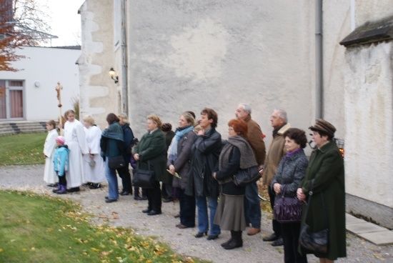 Eine Gruppe von Menschen in Winterkleidung steht in einer Reihe vor einem Gebäude, möglicherweise in Erwartung einer Veranstaltung. Einige tragen Hüte und haben Taschen bei sich.