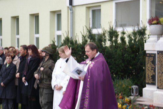Ein Priester in einer violetten Robe winkt einer Menschenmenge zu, die vor einem Gebäude mit einer Gedenktafel steht.
