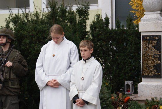 Zwei Kinder in weißen Gewändern stehen neben einem Denkmal, mit einem Soldaten in Uniform im Hintergrund.