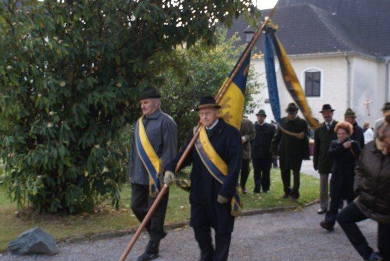 Eine Gruppe von Menschen in traditioneller Kleidung zieht in einer Prozession, mit einer Flagge und einem Stab, vor einem Gebäude.