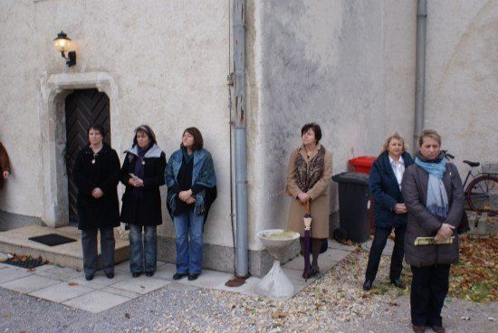 Eine Gruppe von Frauen steht vor einem Gebäude, einige schauen nach oben. Eine hält einen Regenschirm, und ein Fahrrad ist in der Nähe geparkt. Die Szene spielt sich auf einem Kiesbereich mit verstreuten Blättern ab.