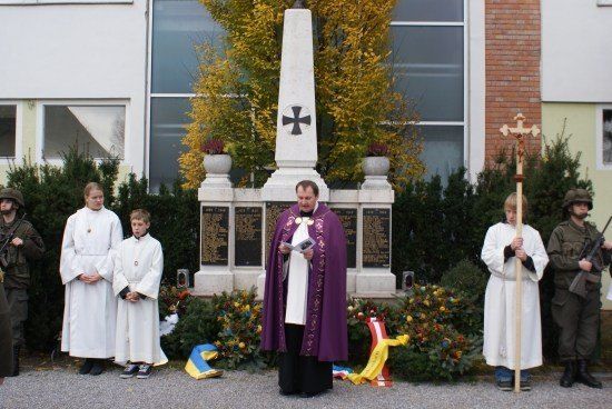Ein Priester in einer violetten Robe steht vor einem Denkmal, mit zwei Kindern in weißen Gewändern und zwei Soldaten in Militäruniformen in der Nähe.