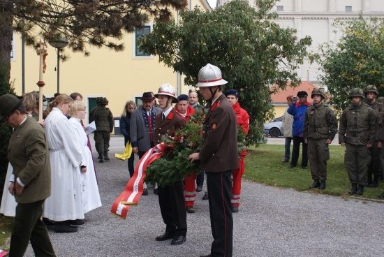 Eine Gruppe von Menschen in Uniformen und Mützen steht um einen Kranz und eine Schleife herum, möglicherweise bei einer Gedenkveranstaltung.