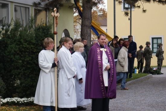 Ein Priester in einer violetten Robe steht mit einem Stab vor einer Gruppe von Menschen in weißen Roben, in der Nähe eines Gebäudes mit einem Baum und Büschen.