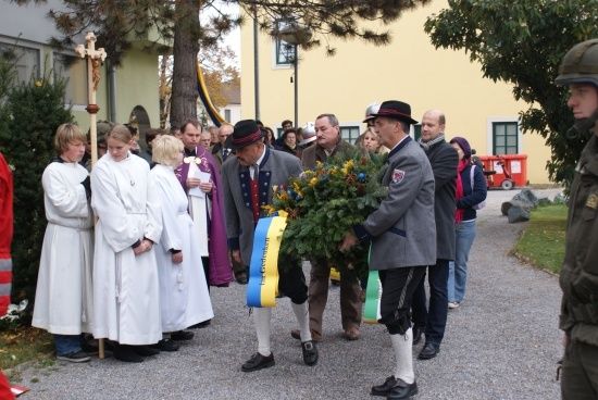 Eine Gruppe von Menschen in traditioneller Kleidung steht in einer Reihe und hält einen Kranz und ein Kreuz. Sie nehmen wahrscheinlich an einer zeremoniellen Veranstaltung teil.