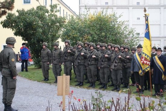 Eine Reihe von Soldaten in Militäruniformen steht in Formation draußen, mit einer Flagge und einem Gebäude im Hintergrund.