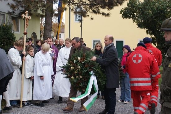 Eine Gruppe von Menschen in weißen Gewändern und einem Mann in einem schwarzen Mantel stehen vor einem Gebäude und halten einen Kranz mit einem grün-weißen Band.