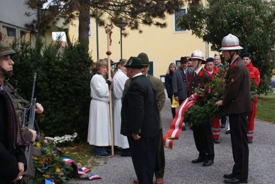 Eine Gruppe von Menschen in Uniformen und Mützen versammelt sich um ein Denkmal, hält eine Girlande und eine Flagge.
