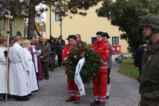 Zwei Feuerwehrleute in roten Uniformen halten einen Kranz vor einer Gruppe von Menschen, einschließlich eines Mannes in einem Militärhelm, der in der Nähe eines Gebäudes steht.