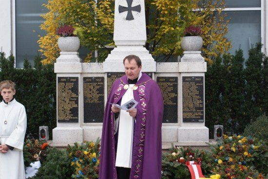 Ein Priester steht vor einem Denkmal und liest aus einem Buch, während ein junger Junge in einem weißen Gewand in der Nähe steht.