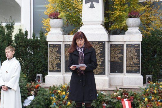 Eine Frau in einem schwarzen Mantel steht vor einem Denkmal und liest ein Papier, während ein junger Junge in einem weißen Gewand in der Nähe steht.