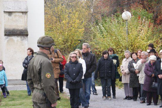 Eine Gruppe von Menschen, darunter ein Mann in Uniform, geht in einer Reihe auf einem Außengelände. Sie sind von Bäumen und einem Gebäude umgeben.