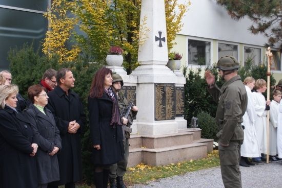 Eine Gruppe von Menschen steht um ein Denkmal mit einem Kreuz oben herum, ein Mann in Uniform salutiert.