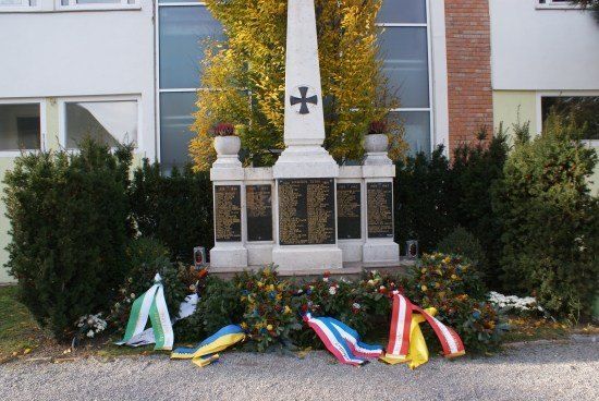 Ein Denkmal mit einem Kreuz oben, umgeben von Blumen und Bändern, vor einem Gebäude mit Glasfenstern.