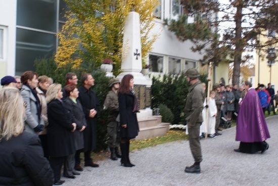 Eine Gruppe von Menschen in Winterkleidung steht vor einem Gebäude mit einem weißen Denkmal und einem Kreuz. Eine Person in Militäruniform steht rechts.