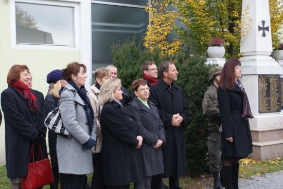 Eine Gruppe von Menschen in Winterkleidung steht vor einem Denkmal mit einem Kreuz und einer Tafel.