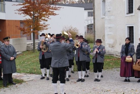 Eine Blaskapelle in traditionellen Uniformen steht vor einem Gebäude, wobei ein Mitglied führt und andere Instrumente spielen.