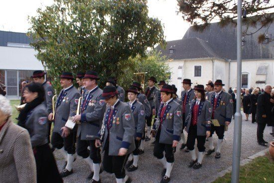 Eine Gruppe von Menschen in traditionellen Uniformen und Hüten marschiert in einer Reihe, wahrscheinlich für eine Parade. Sie tragen Musikinstrumente. Hinter ihnen stehen mehrere Menschen in der Nähe eines Gebäudes.