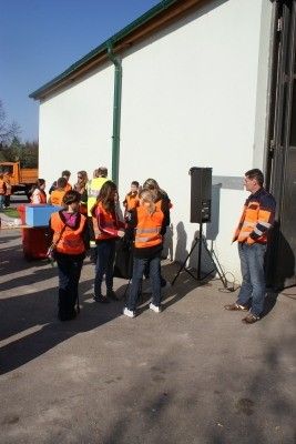 Eine Gruppe von Menschen in orangenen Westen steht vor einem Gebäude mit einem Lautsprecher und einem Mann, der mit ihnen spricht.