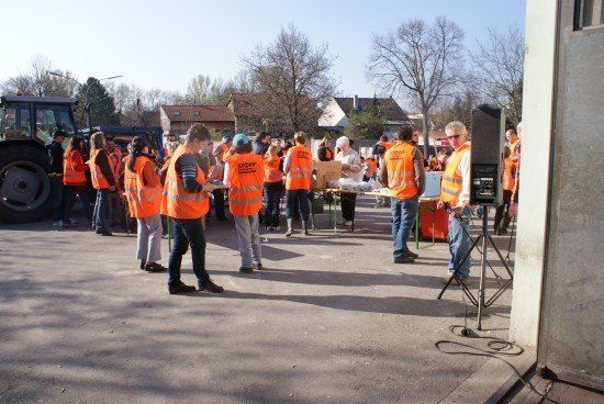 Eine Gruppe von Menschen in orangefarbenen Westen steht in einer Reihe, wahrscheinlich auf etwas wartend. Ein Mann mit einem weißen Hut steht vor einem Tisch mit Gegenständen darauf. Ein Traktor ist hinter ihnen geparkt.
