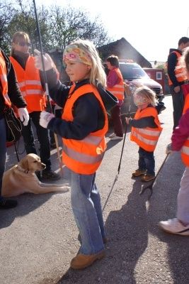 Ein junges Mädchen in einer orangefarbenen Sicherheitsweste, die einen Stock hält, steht mit einem Hund und einem kleinen Mädchen in ähnlichen Westen, während andere in orangefarbenen Westen hinter ihnen stehen.