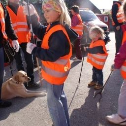Ein junges Mädchen in einer orangefarbenen Sicherheitsweste, die einen Stock hält, steht mit einem Hund und einem kleinen Mädchen in ähnlichen Westen, während andere in orangefarbenen Westen hinter ihnen stehen.