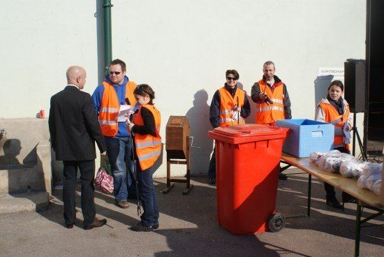 Eine Gruppe von Menschen in orangefarbenen Westen steht draußen in der Nähe eines Tisches mit Tüten und einem roten Mülleimer. Ein Mann in Anzug spricht mit einer Frau, die ein Papier hält.