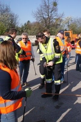 Eine Gruppe von Personen in Sicherheitswesten versammelt sich auf einer Straße, wobei einer der Männer sich bücken und ein anderer einen Stock hält. Sie scheinen eine Outdoor-Aktivität oder Inspektion durchzuführen.