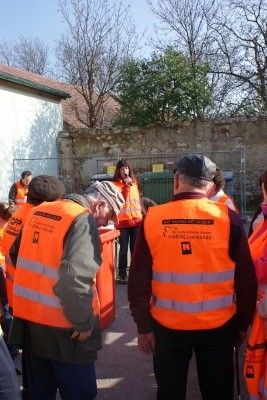 Eine Gruppe von Menschen in reflektierenden orangefarbenen Westen steht vor einem Gebäude. Einige schauen nach unten, während andere in Gespräche vertieft sind. Eine Frau in einem roten Shirt steht in der Nähe eines grünen Tores. Bäume und eine Mauer sind im Hintergrund.