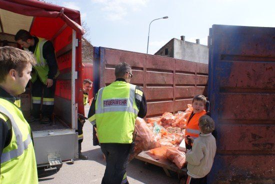 Eine Gruppe von Menschen, darunter Feuerwehrleute, laden orangefarbene Säcke in einen Lastwagen. Ein Kind beobachtet.